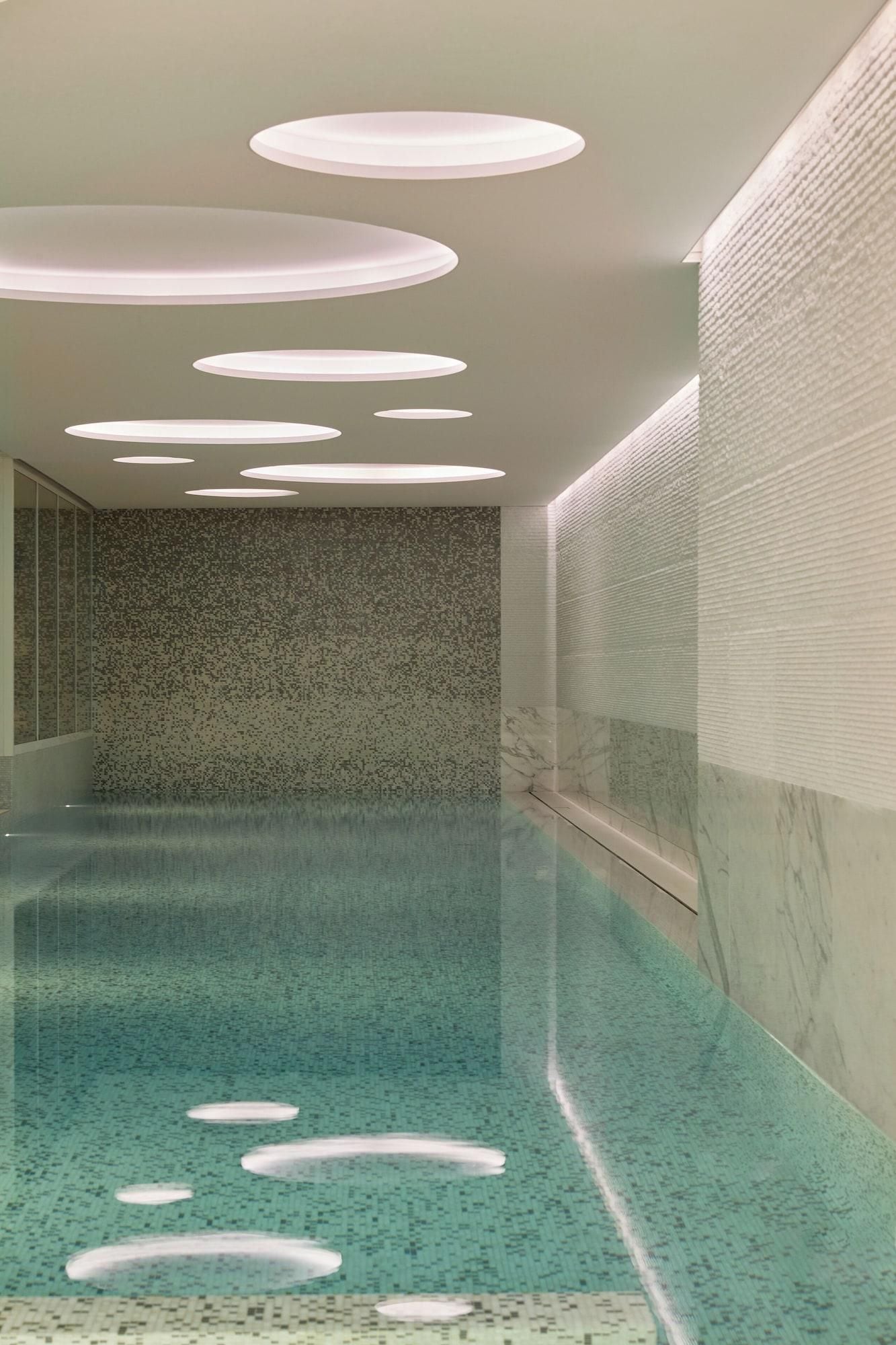 A modern indoor swimming pool with a clear blue water surface, a textured wall with a speckled pattern, and circular ceiling lights.