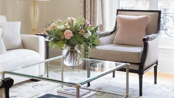 Uma sala de estar com uma mesa de centro de vidro, um sofá bege, uma poltrona escura e um vaso com flores.