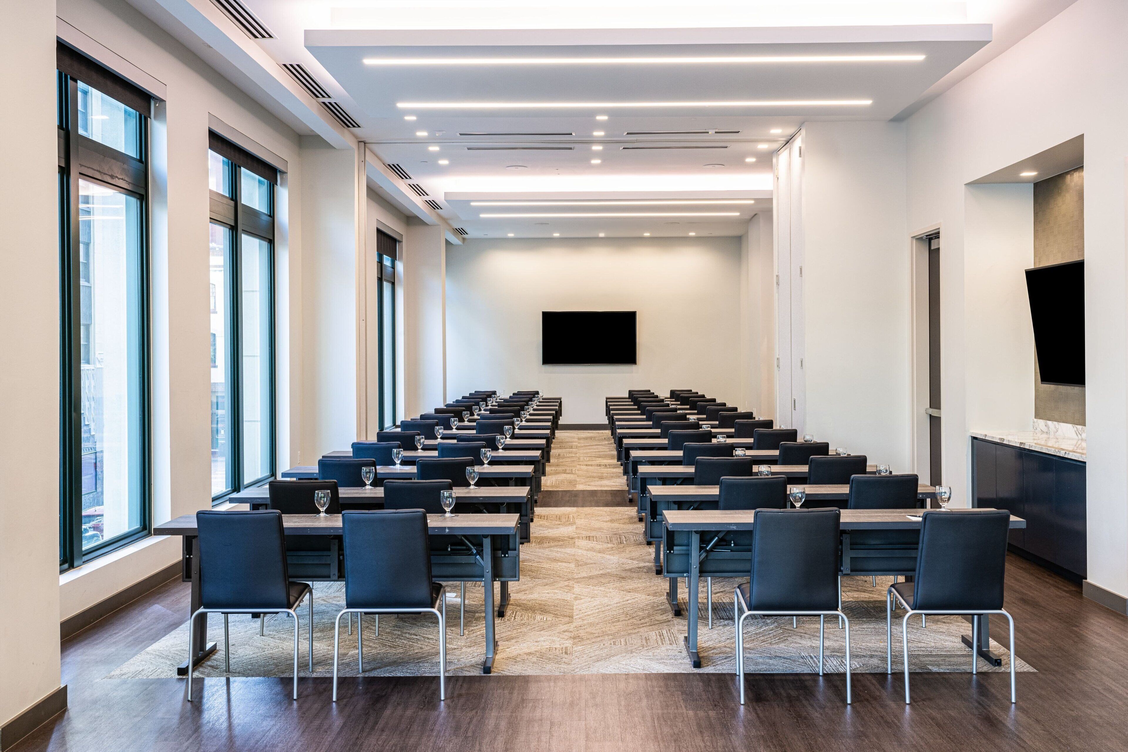 A conference room with rows of tables and chairs, a large flat-screen TV, and a kitchen area.