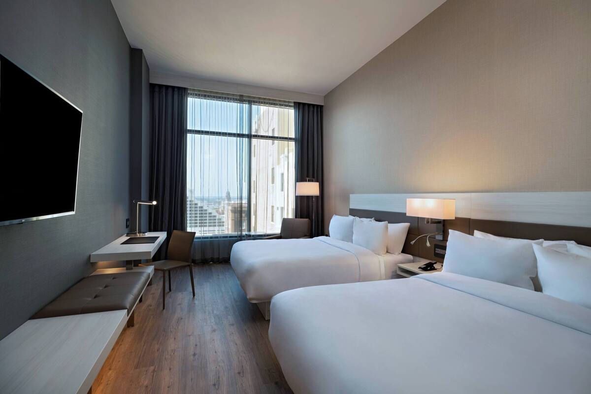 A hotel room with two beds, a desk, a chair, a TV, and a window with curtains.
