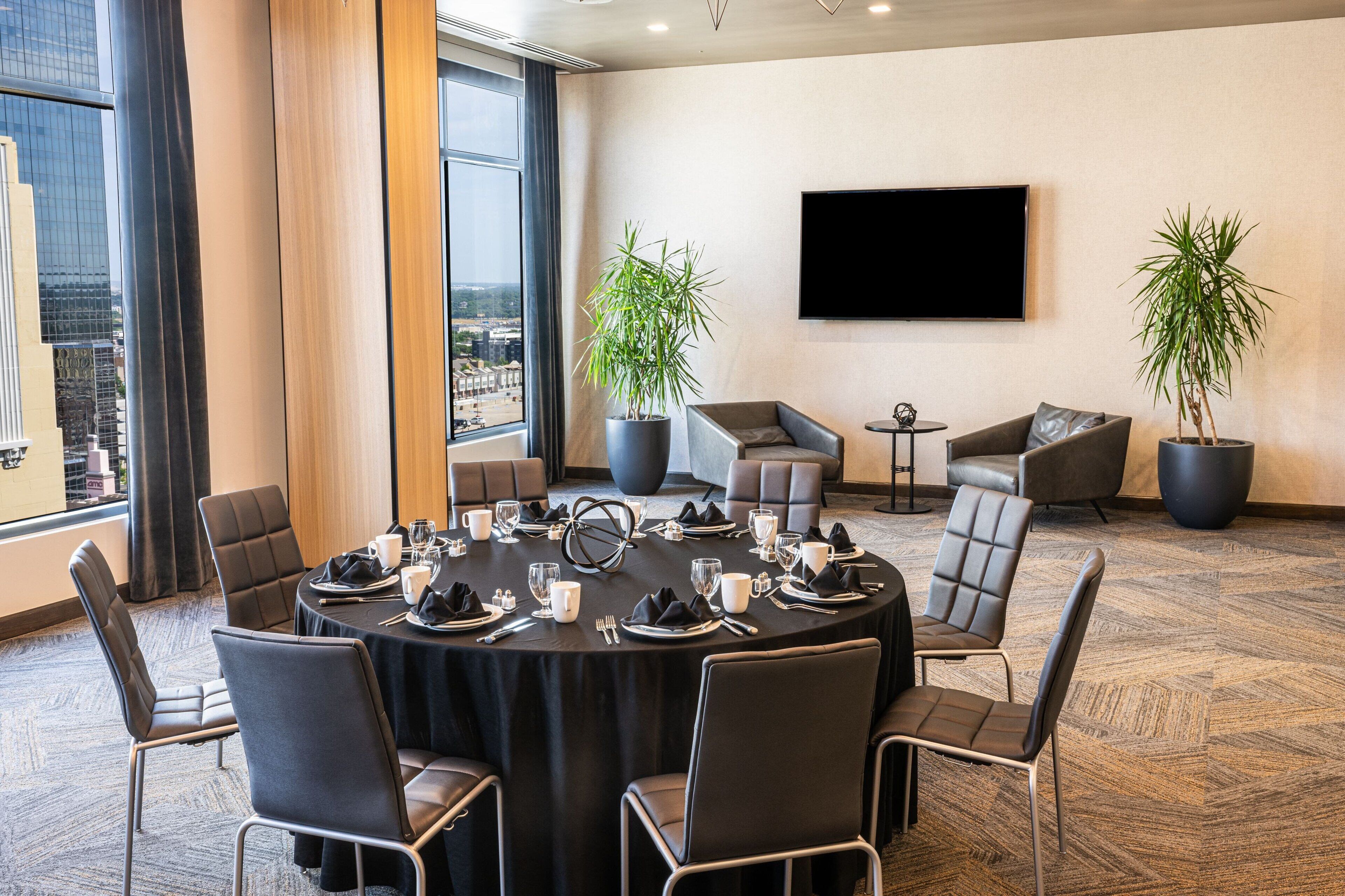 A conference room with a round table set for a meeting, surrounded by chairs, with a large potted plant and a flat-screen TV on the wall.