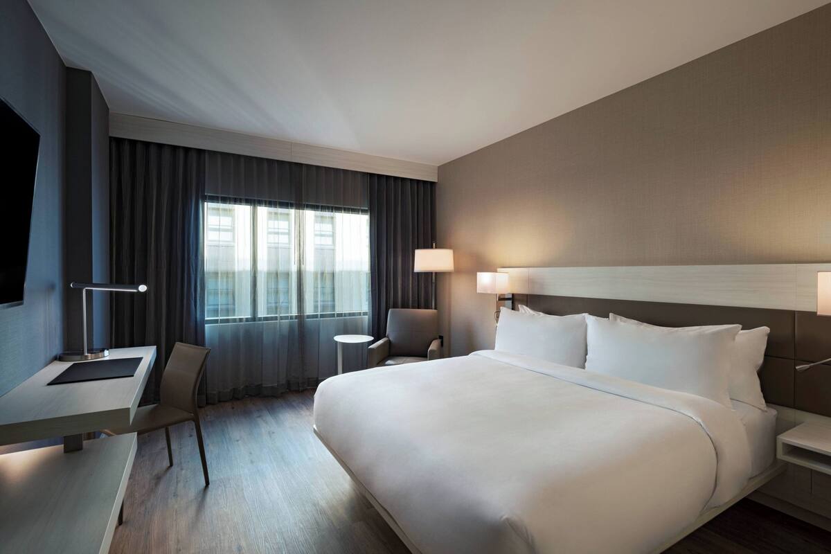 A hotel room with a large bed, a desk, a chair, a lamp, and a TV.