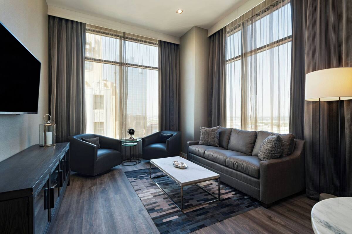 A modern hotel room with a sofa, a coffee table, a TV, and large windows with curtains.