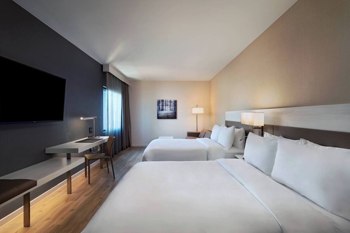 A hotel room with two beds, a desk, a TV, and a painting on the wall.