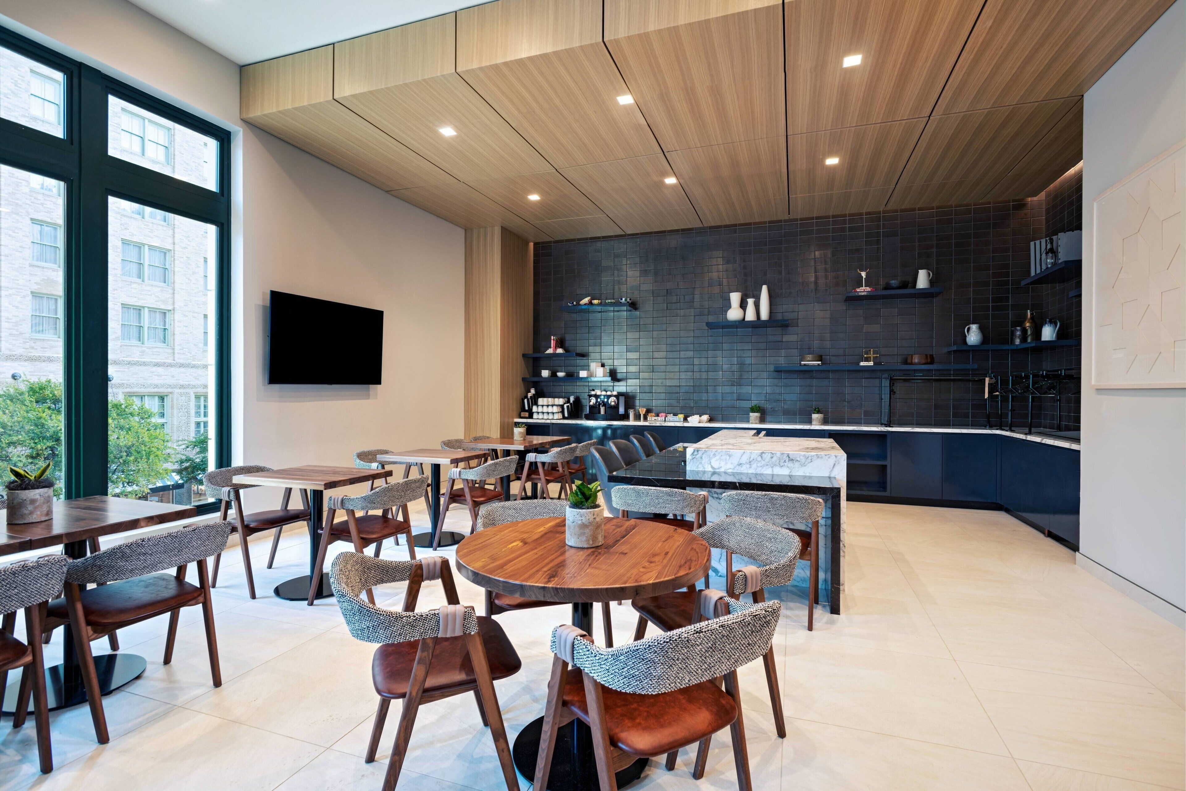 A modern dining area with round tables and wicker chairs, a kitchen with dark tiles and wooden cabinets, and a large window offering a view of buildings outside.