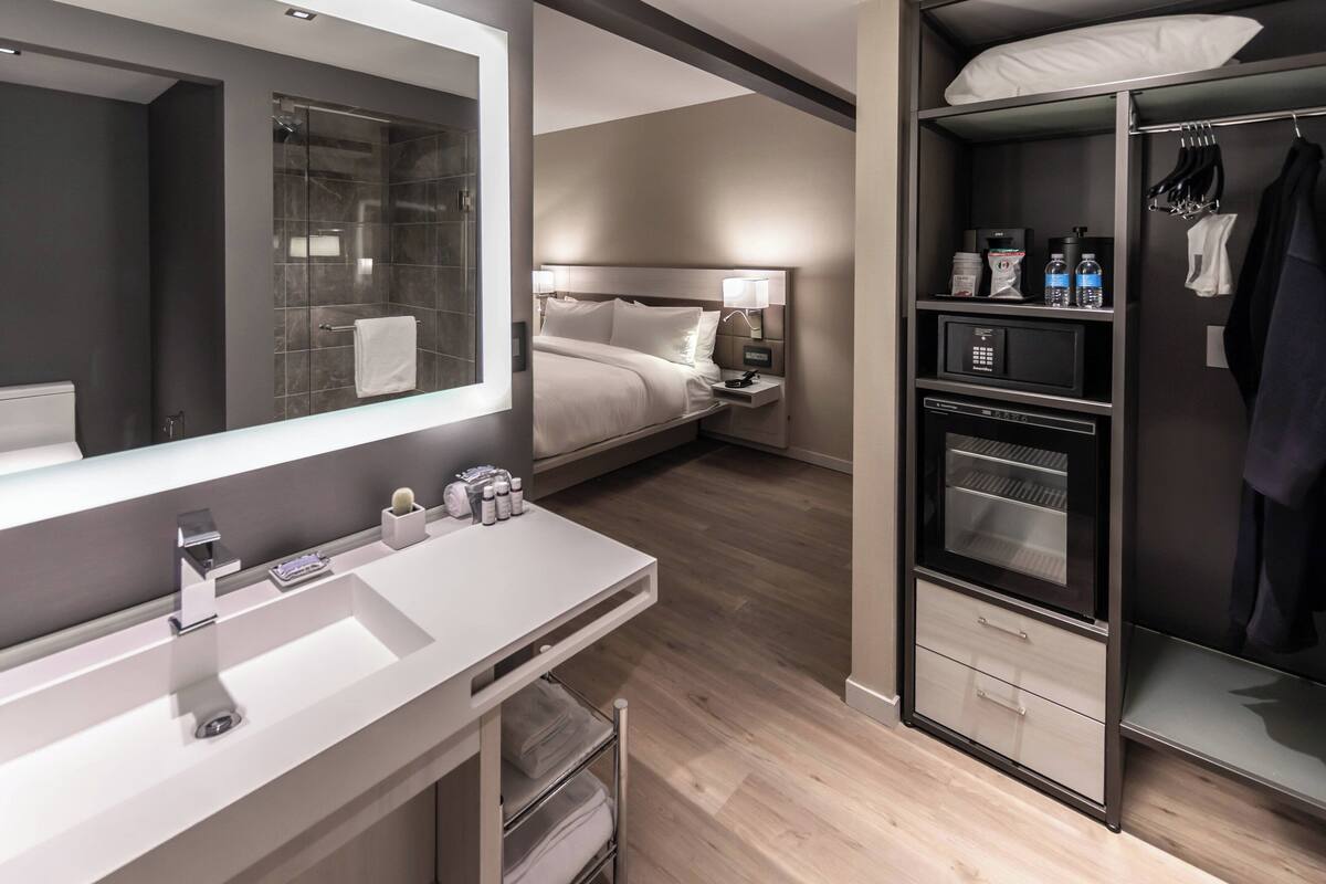 A modern hotel room with a large mirror, a white sink, a bed with a headboard, a nightstand with a lamp, a wardrobe, and a built-in microwave.