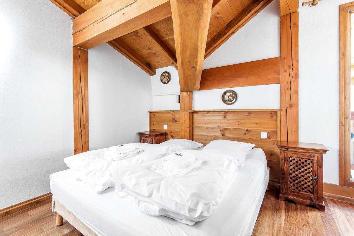Un dormitorio con techo de madera, cama con ropa de cama blanca, mesitas de noche de madera y cabecera de madera.