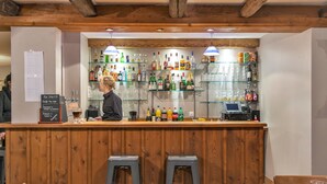 Zona de bar con mostrador y taburetes de madera, estantes con diferentes botellas y vasos, y una pizarra de menú.
