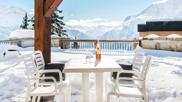 Un área de estar al aire libre cubierta de nieve, con una mesa y sillas, con vistas a un paisaje montañoso.