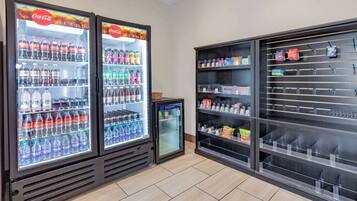 Uma área de máquinas automáticas com refrigeradores de Coca-Cola, um bebedouro de água Dasani e uma prateleira com diversos snacks e artigos de higiene pessoal.