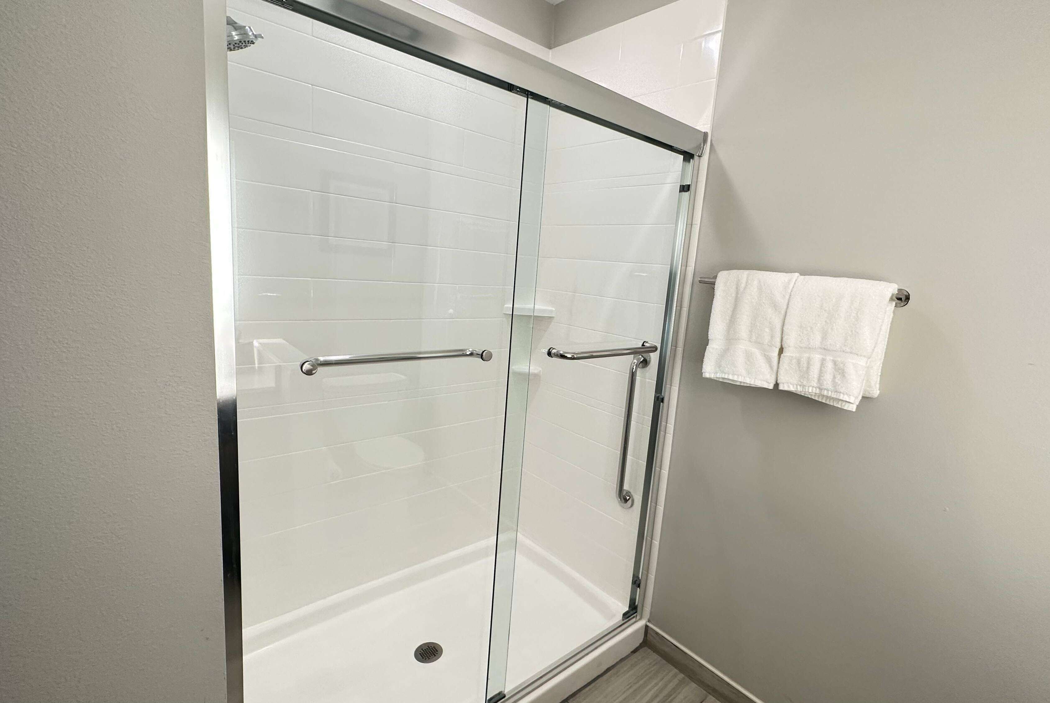 A glass-enclosed shower with a white tiled wall, a metal handle, and a white towel hanging on a hook.