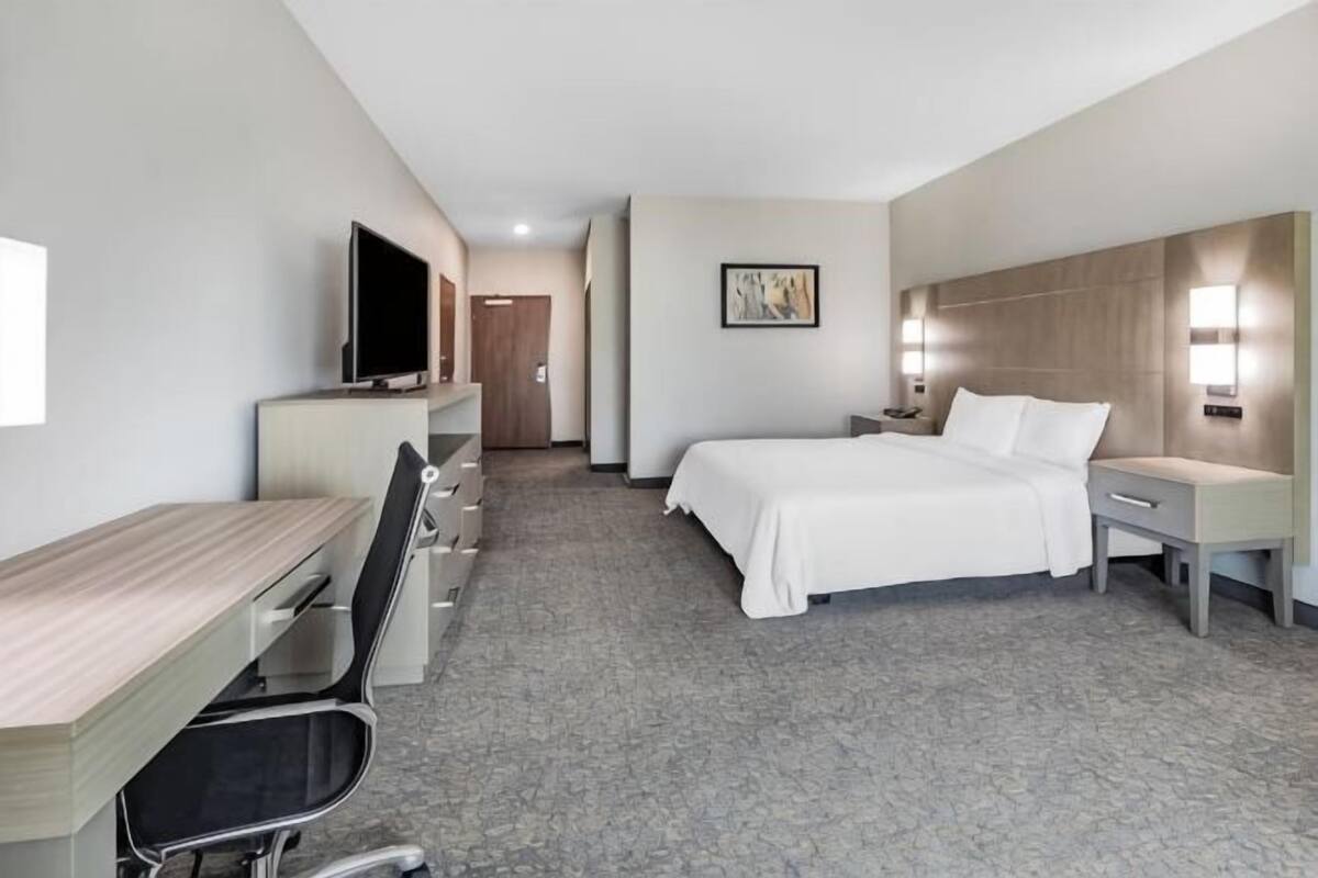 A hotel room with a bed, desk, chair, TV, and artwork on the wall.