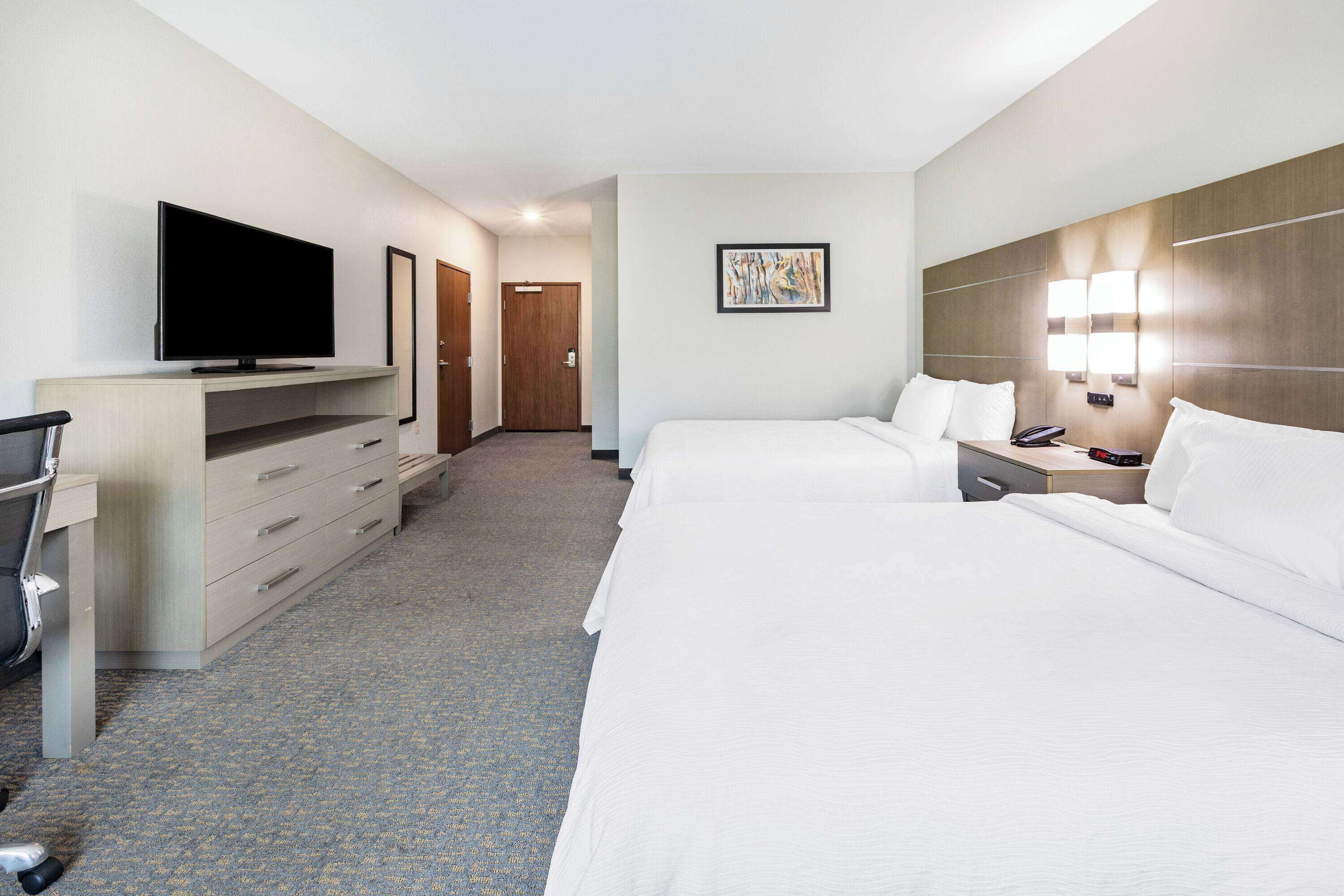 A hotel room with a TV on a desk, two beds, a desk with drawers, a chair, and a door in the background.