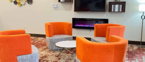 A modern lounge area with orange and grey upholstered chairs, a flat-screen TV, and a fireplace.