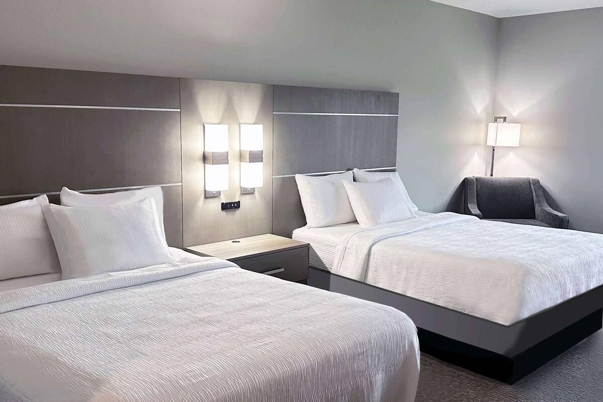 A hotel room with two beds, a nightstand, a lamp, and a chair.