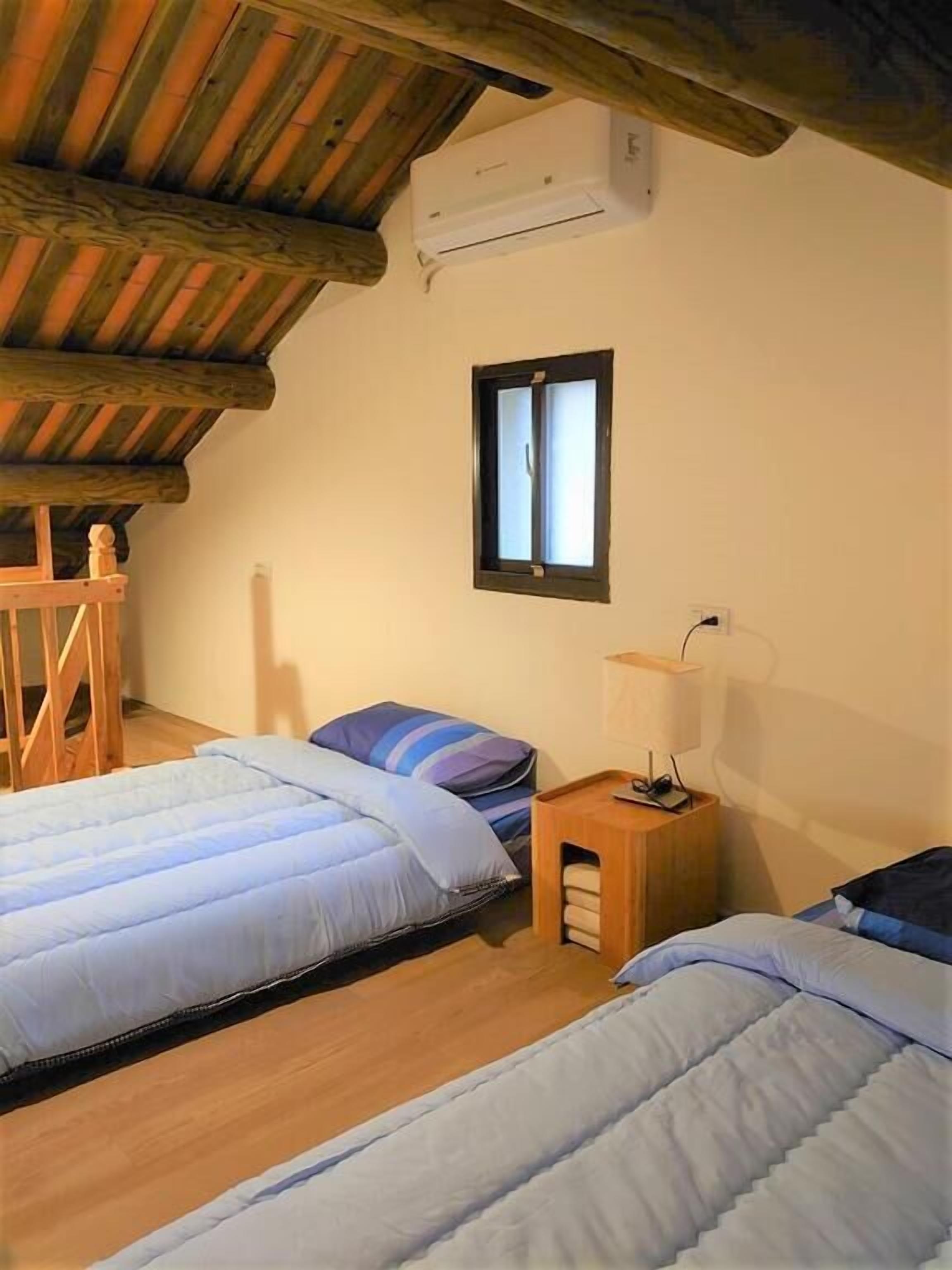 A cozy room with two beds, a wooden bench, a small table with a lamp, and a window with blinds.
