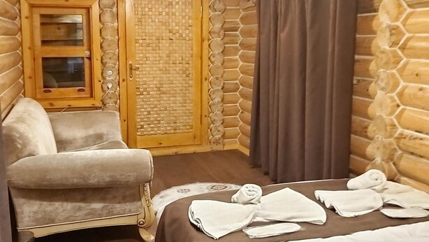 A cozy log cabin room with a bed, a chair, a small table with towels, a wooden door, and a window.