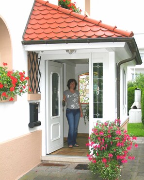 A woman standing at the entrance of a house with a red tiled roof, white walls, and a glass door.
