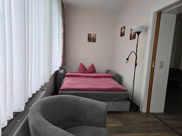 A bedroom with a bed, a chair, a standing lamp, and a door leading to another room.