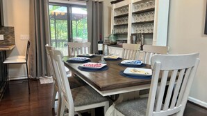 Un comedor con una mesa de madera dispuesta para cuatro personas, rodeada de sillas blancas con cojines grises. Una botella de vino y copas están sobre la mesa, y se observa una estantería con adornos en el fondo.