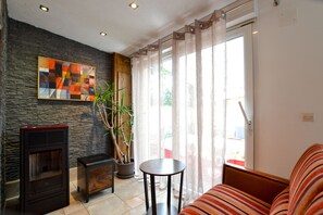 A living room with a stone wall, a fireplace, a sofa, a table, and a large window with curtains. - Rural Cottage, indoor swinming pool and spa, playg (Ostheim)