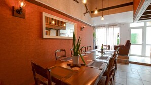 A dining area with a long wooden table, chairs, a potted plant, and a mirror on the wall.