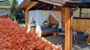 Une terrasse en bois avec un lit à baldaquin, un coin salon et le tronc d’un arbre.