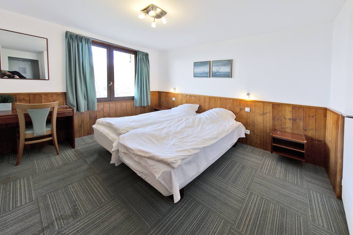 Habitación de hotel con cama doble, escritorio con silla, mesita de noche, ventana con cortinas y dos cuadros en la pared.