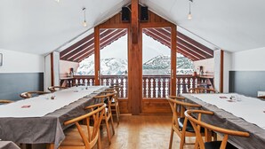 Een eetruimte met houten tafels en stoelen, grote ramen met uitzicht op besneeuwde bergtoppen, en een houten balkenplafond.