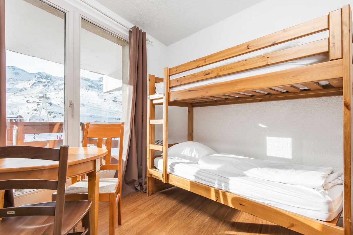 Ein Schlafsaalzimmer mit Etagenbetten, einem Holztisch und Stühlen, einem Fenster mit Vorhängen und Blick auf eine verschneite Landschaft.