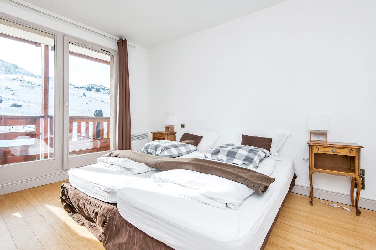 Ein Schlafzimmer mit einem großen Bett, einem Nachttisch aus Holz, einem Fenster mit Blick auf eine verschneite Landschaft und einem Balkon.