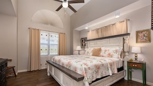 Un dormitorio con una cama grande, piso de madera, ventilador de techo y ventana con cortinas.