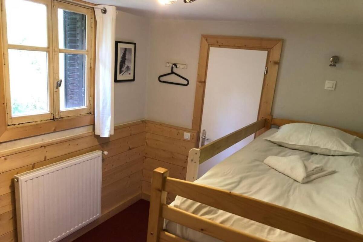 Ein kleines, gemütliches Zimmer mit einem hölzernen Bett, einem Heizkörper, einem Fenster mit Vorhängen und einer Holztür.