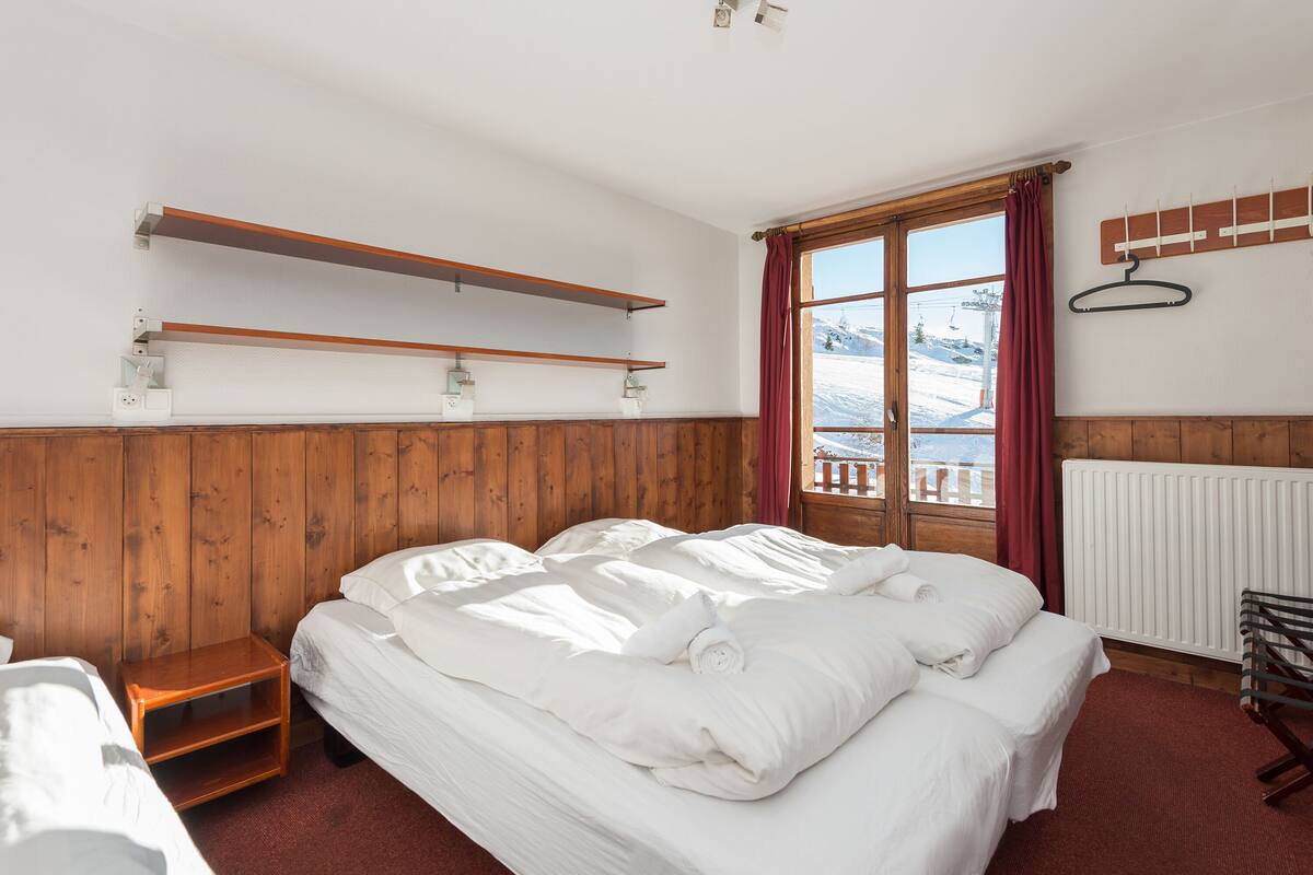 Ein Schlafzimmer mit Holz-Kopfteil, einem großen Bett mit weißer Bettwäsche, einem Nachttisch aus Holz, einem Fenster mit Blick auf eine verschneite Landschaft und einer Heizung.