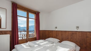 A room with two beds, wooden paneling, a window with a view of mountains, and a radiator. - Klubhotel Le Refuge by Skinetworks (Huez)