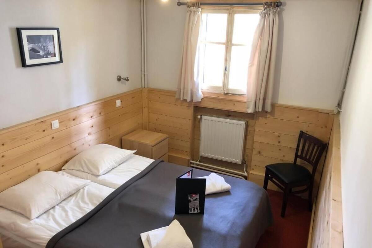 Ein Zimmer mit Holzwänden, einem Bett, einem Sessel, einem Heizkörper und einem Fenster mit Vorhängen.