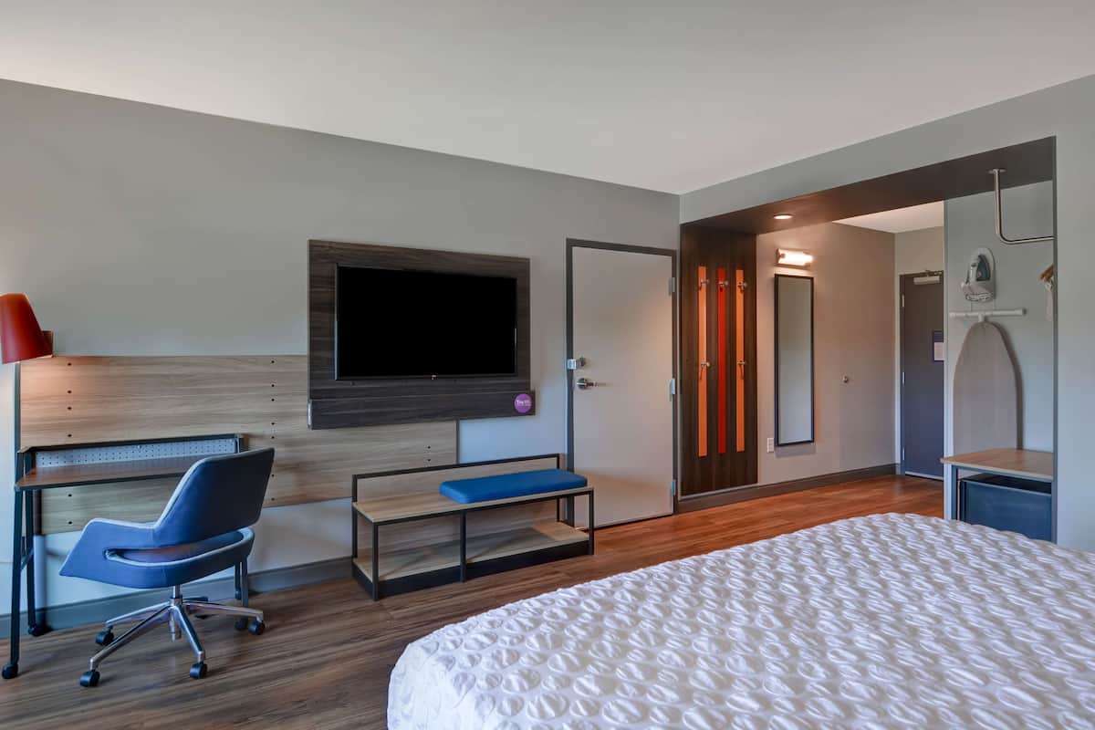 A modern hotel room with a large bed, a desk with a chair, a TV mounted on the wall, and a bathroom visible through an open door.