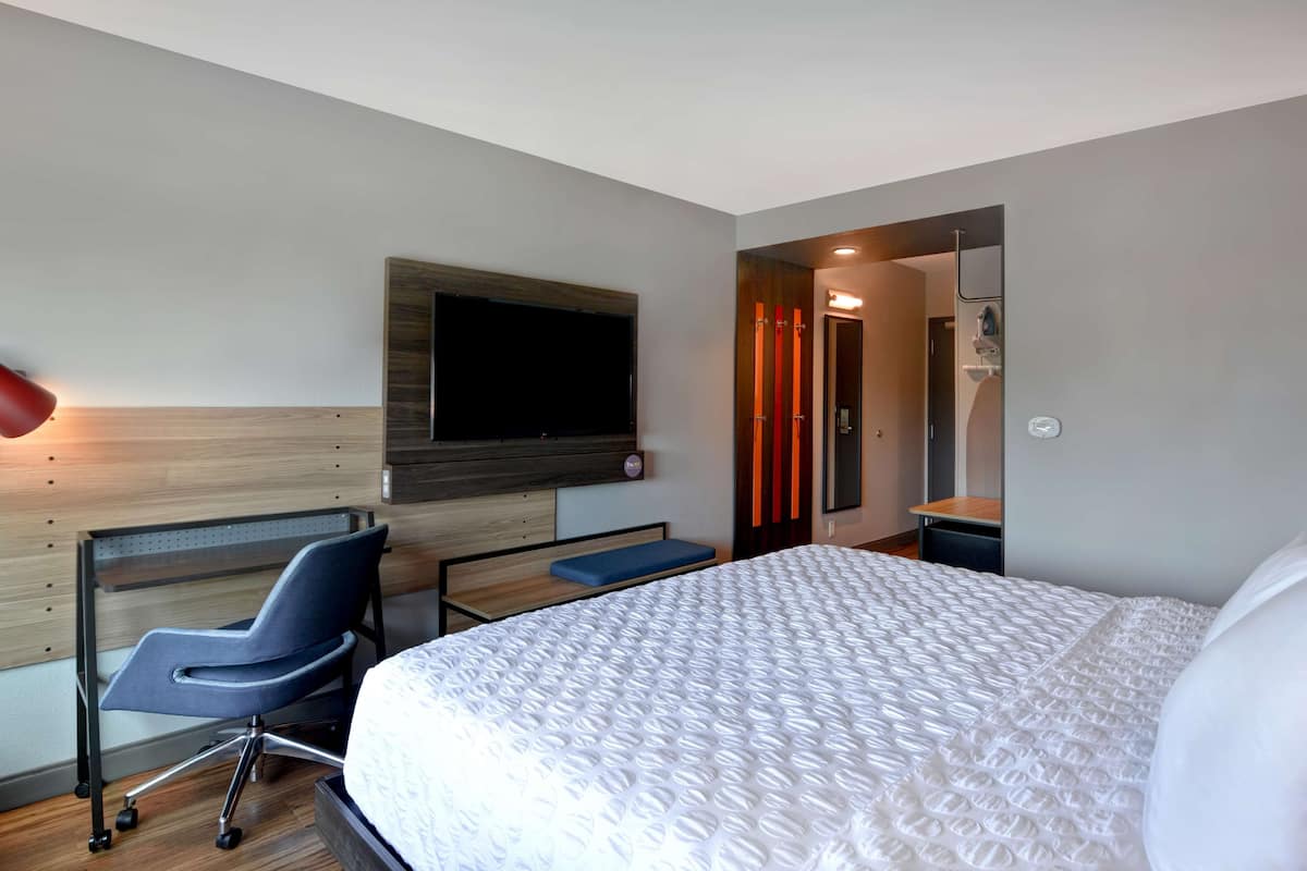 A modern hotel room with a large bed, a desk with a chair, a flat-screen TV mounted on the wall, and a shower area visible through a glass door.