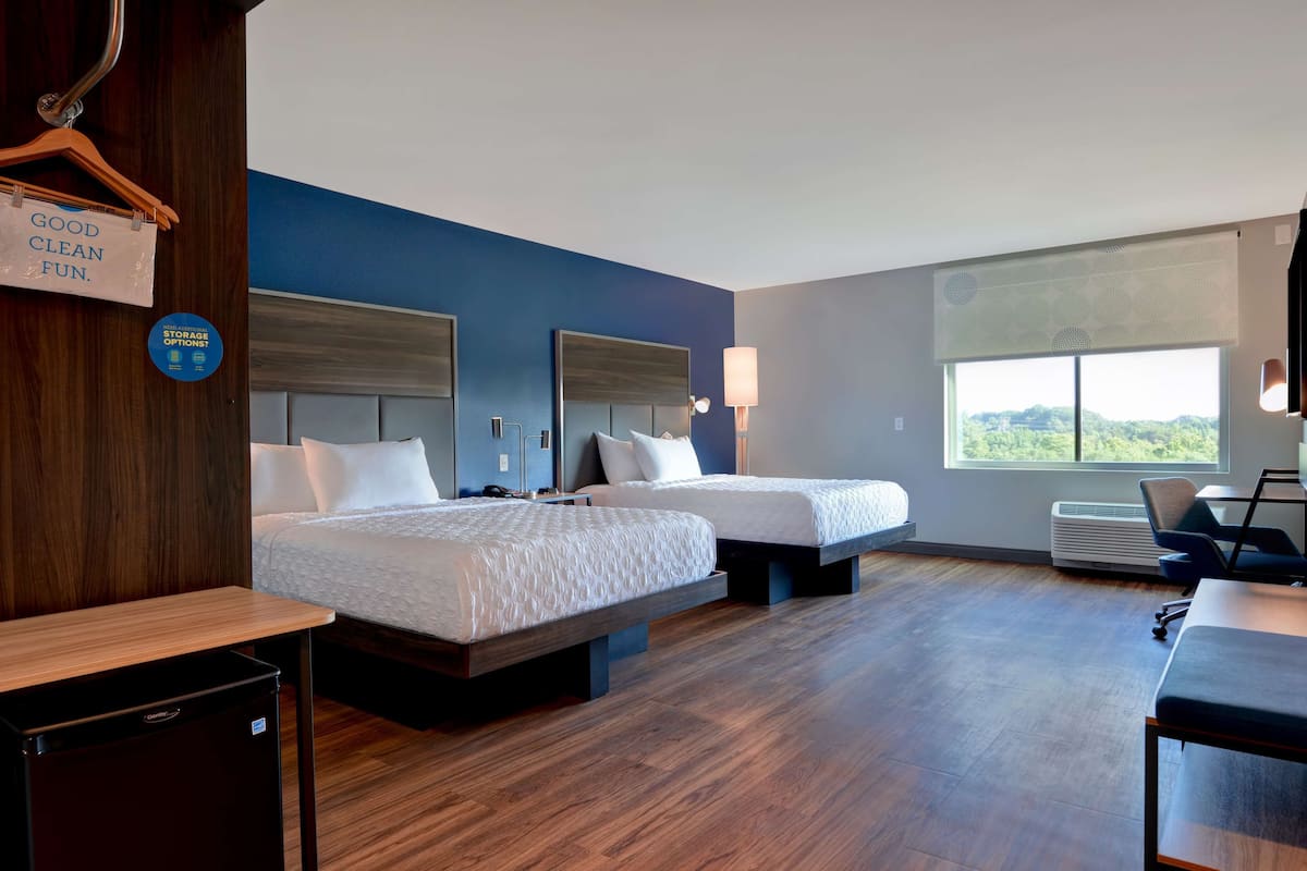 A hotel room with two beds, a desk, a chair, a small fridge, a window with a view, and a sign indicating storage options.