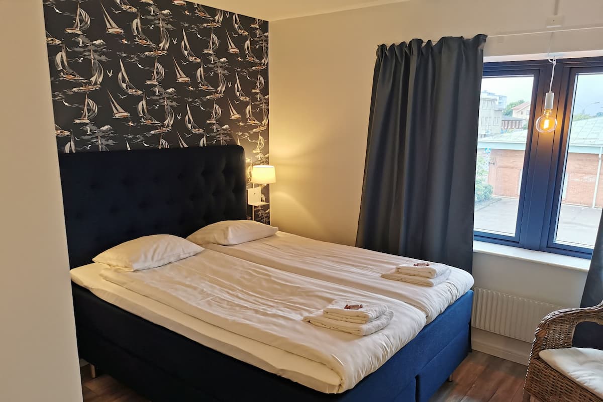 Uma cama bem arrumada com almofadas, cabeceira, luminária e janela com cortinas.