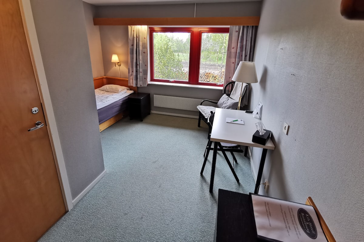 Quarto de hotel compacto com uma cama de solteiro, uma pequena escrivaninha com cadeira, uma janela com cortinas e um radiador.