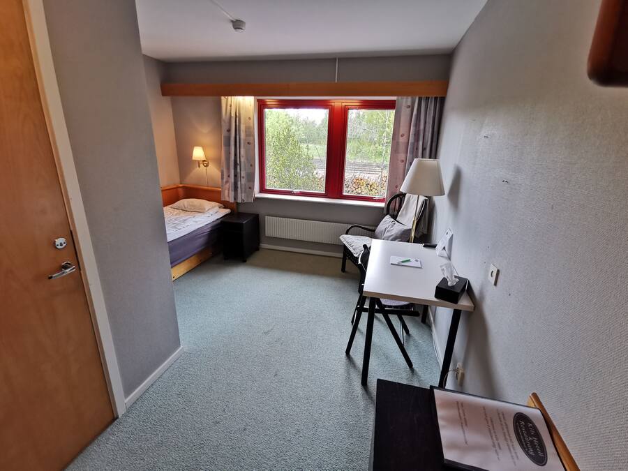 A compact hotel room with a single bed, a small desk with a chair, a window with curtains, and a radiator.