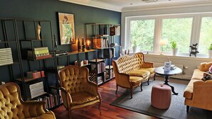 A cozy living room with a dark blue wall, wooden flooring, and a large window with a view of greenery. There are two armchairs, a sofa, and a coffee table.