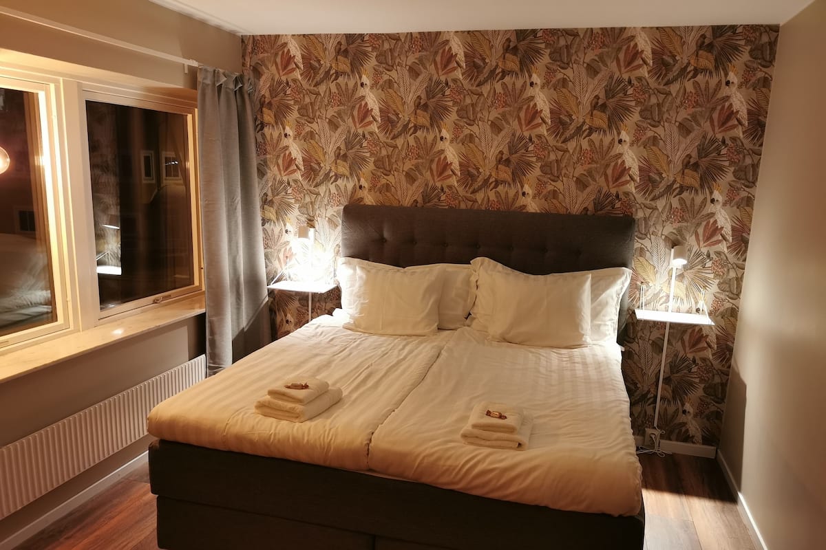 Cama bem arrumada com lençóis e toalhas brancas, cabeceira escura e papel de parede com padrão floral.