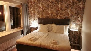 A neatly made bed with white linens and towels, a dark headboard, and a floral wallpaper pattern.