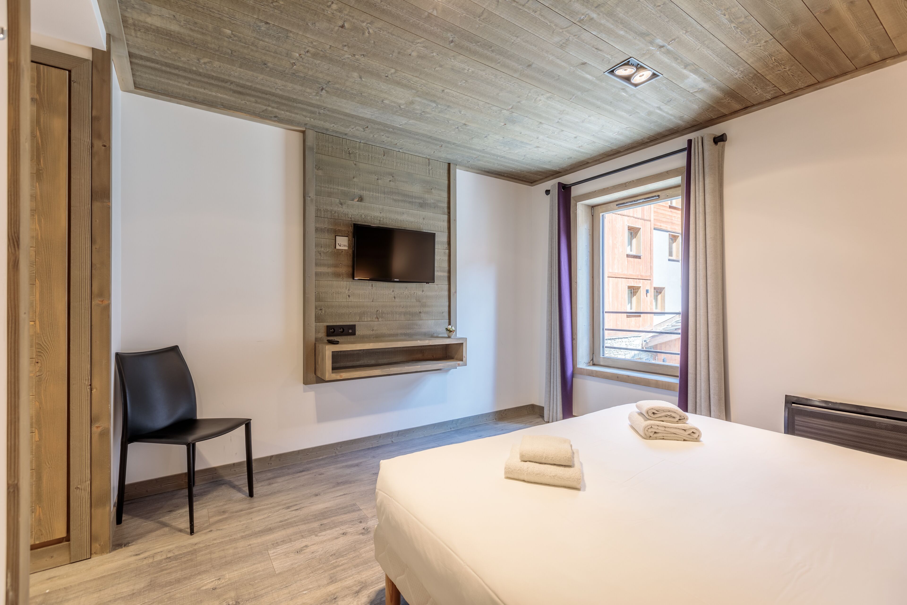 Une chambre d’hôtel avec un plafond en bois, un téléviseur à écran plat fixé au mur, un lit avec des serviettes soigneusement pliées, une chaise et une fenêtre avec des rideaux.