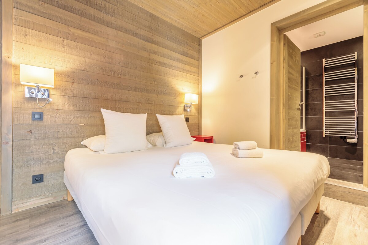 Habitación de hotel con pared de madera, una cama grande con ropa blanca, dos lámparas de noche y un baño visible con perchero.