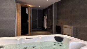A modern bathroom with a hot tub, dark tiled walls, and recessed ceiling lights.