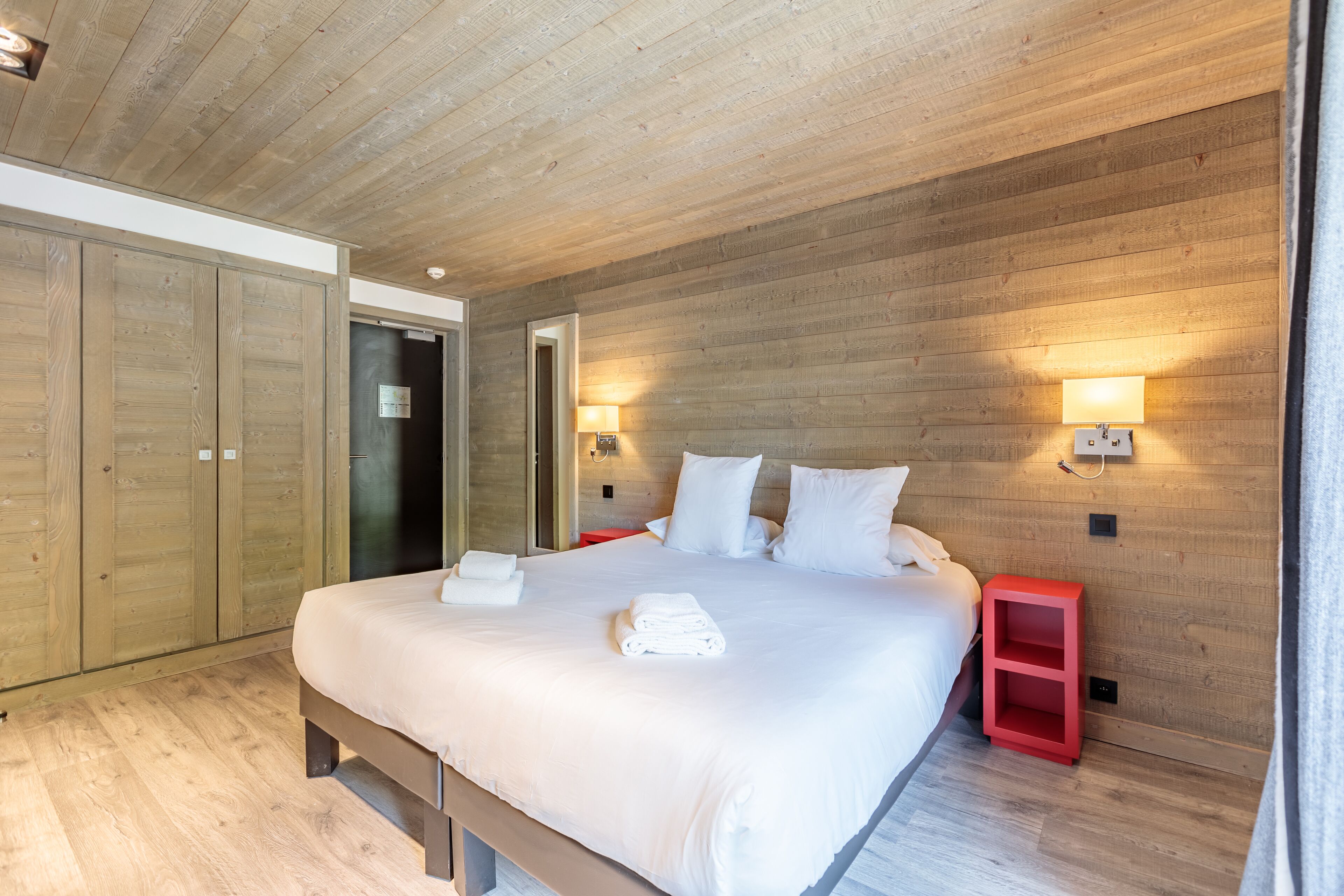 A modern bedroom with a wooden ceiling, a large bed with white linens, two bedside lamps, and a red nightstand.