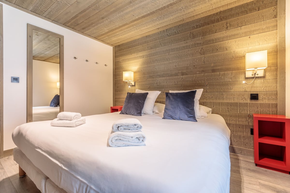A neatly made bed with white linens and folded towels, a wooden headboard, and two wall-mounted reading lights.